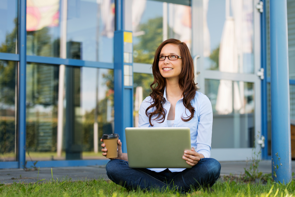 Image of a young woman smiling on something, while holding coffee and laptop, sitting crosslegged in the lawn.